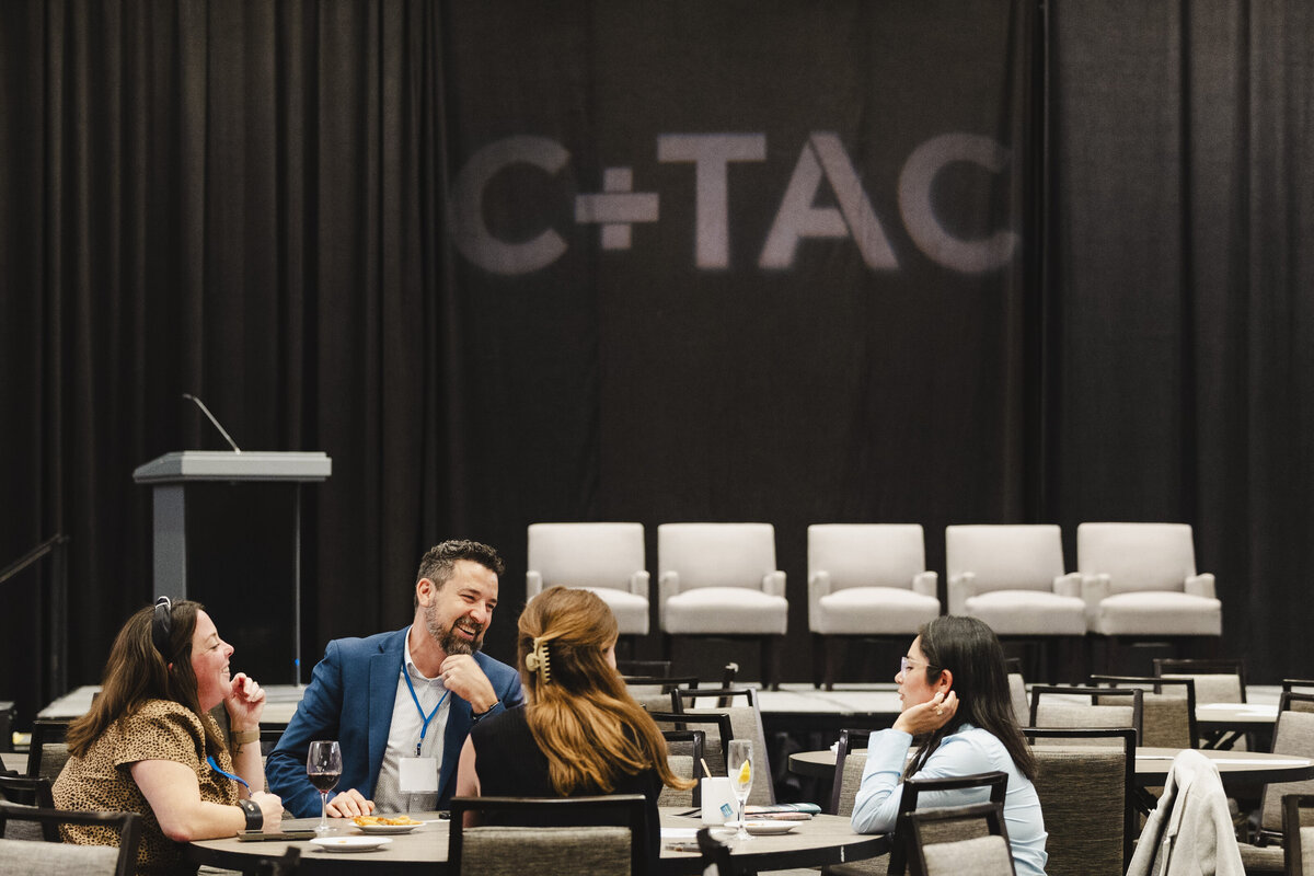 People discuss at a table with an event sign in the back that says C+TAC.