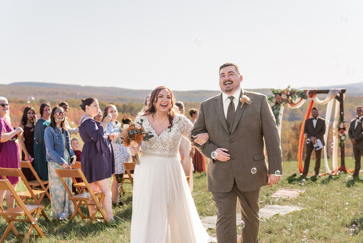 Bride and groom walk back down the aisle laughing after their outdoor fall wedding ceremony near Pittsburgh, surrounded by guests blowing bubbles - photographed by Bree Stewart of LLP.