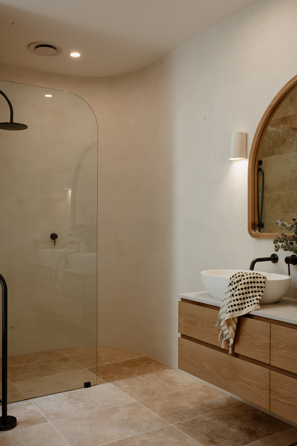 Minimalist bathroom with a curved glass shower, wooden vanity, round mirror, and beige tiles, expertly crafted by a luxury home design team in Newcastle.
