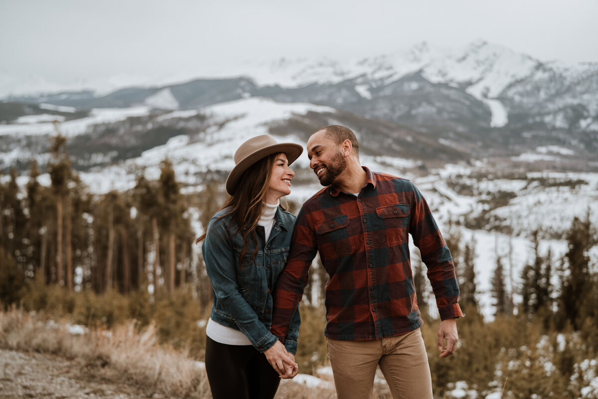 Engaged couple holds hands and smiles with snowy Breckenridge mountains behind them.