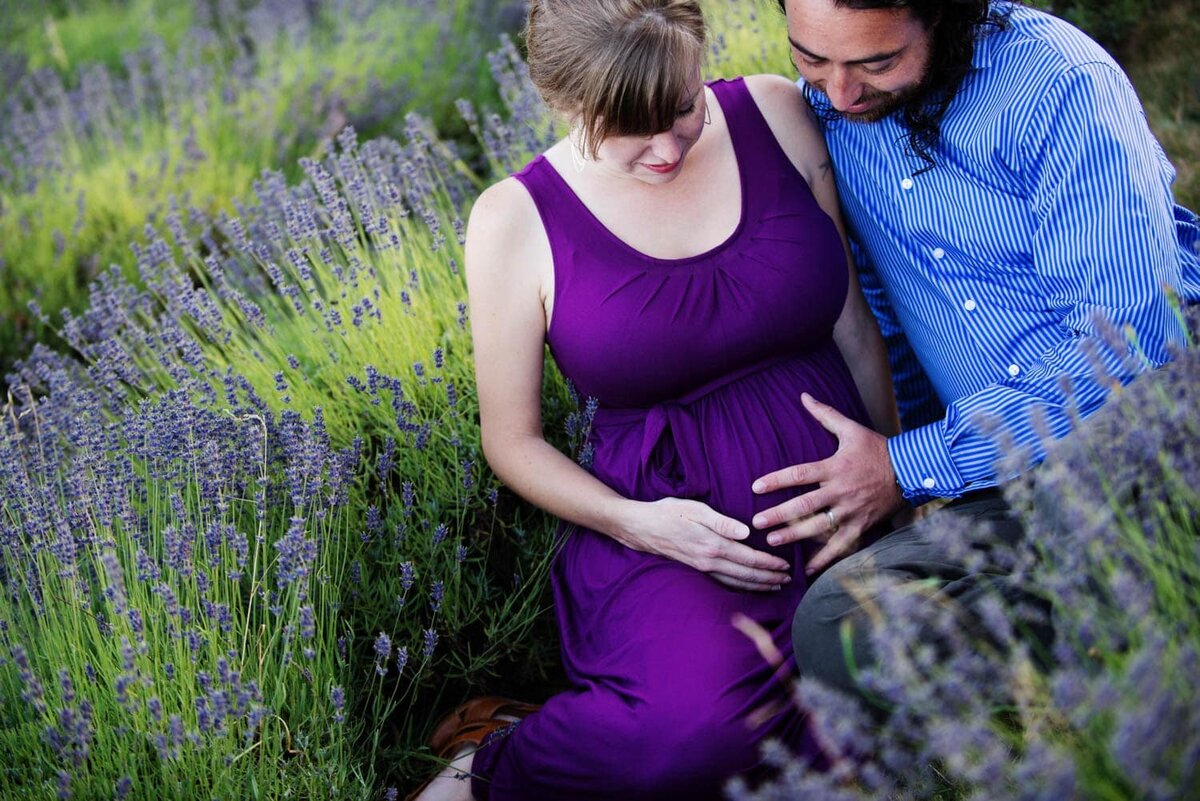Colorful expecting mom in a purple dress and dad in a blue dress shirt , sitting together, surrounded by lavender flowers.