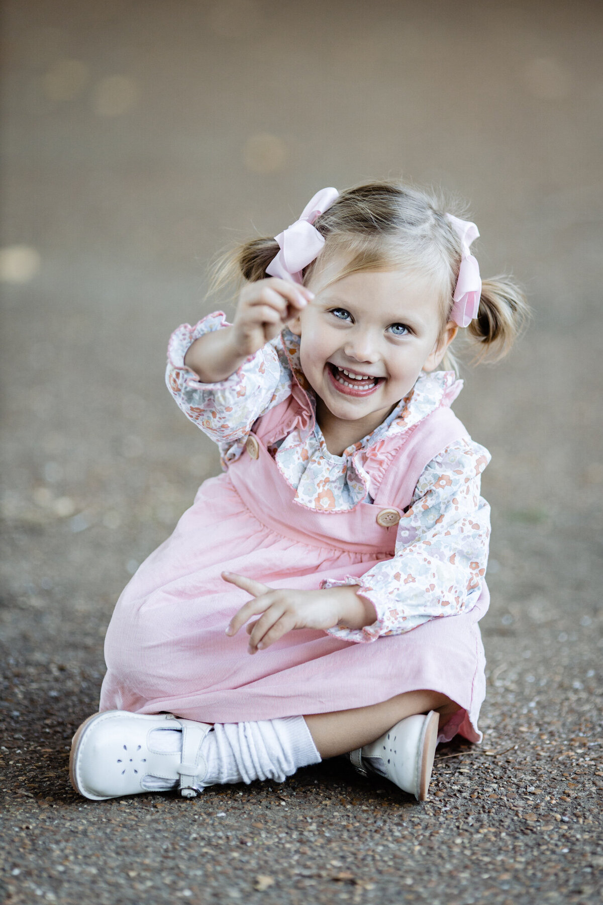 A close up of a little girl in a pink dress sitting on the pavement. She's holding up a rock to show the camera.