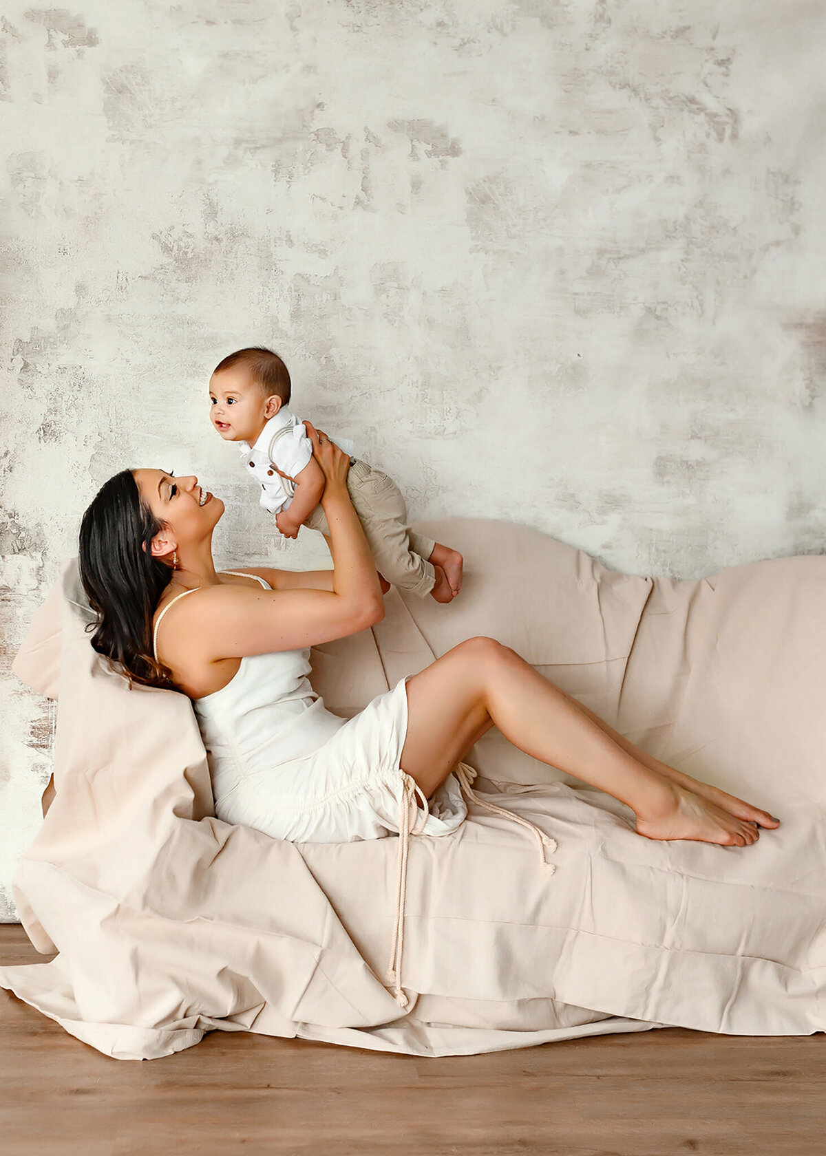 mom holding her son up and lovingly looking at him while sitting on a canvas covered couch against a white textured background