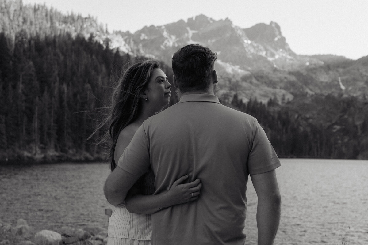 Black-and-white cinematic photograph of an engaged couple embracing by a serene mountain lake during their intimate documentary-style engagement session.