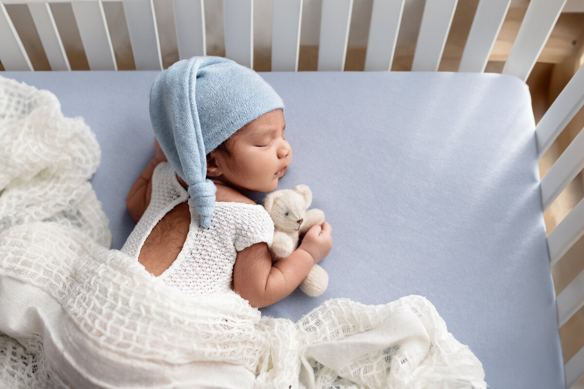 Newborn baby in a blue bonnet lying in a crib with white knitted blankets.