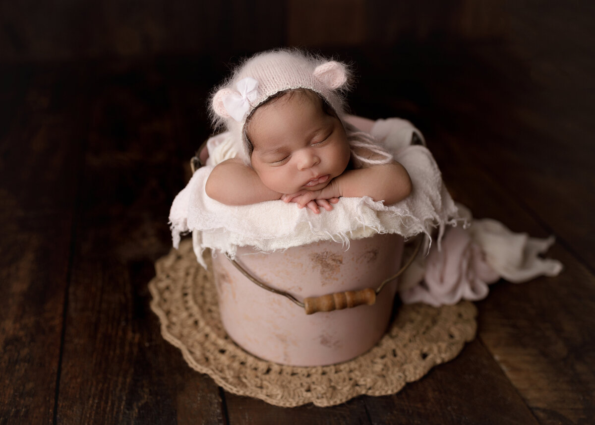 Baby girl sleeping in a blush bucket for studio newborn  photo session. Baby's hans are folded under her chin and she is resting her cheek on her left arm. baby is wearing a fuzzy knit teddy bear bonnet.