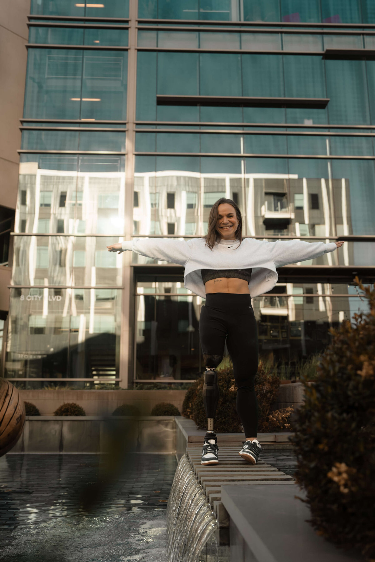 An adaptive athlete stands confidently with arms outstretched on a decorative urban waterfall ledge, wearing Nike athletic wear including a white cropped sweatshirt and black leggings. She is smiling warmly, with a prosthetic leg visible, against the backdrop of a modern glass building.