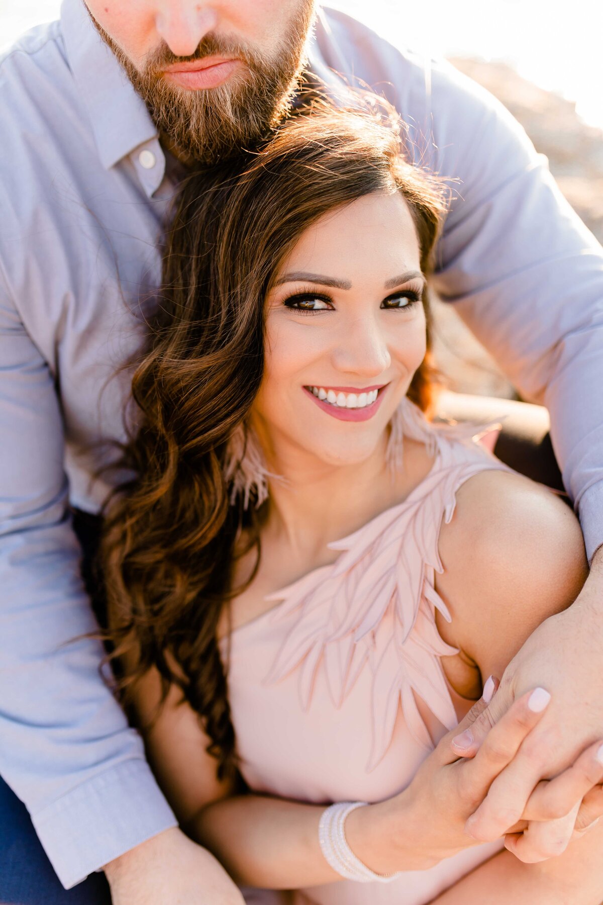 Bride to be smiles at the camera as she nestles in her mans arms. Light and Airy Luxury Photography.