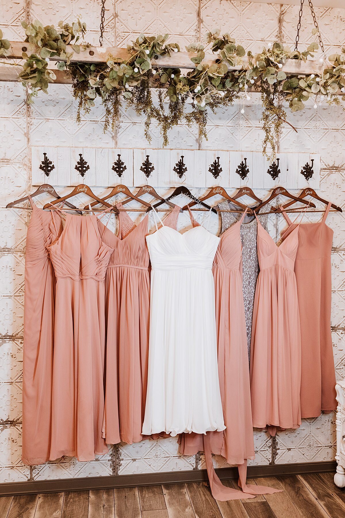 Mismatched blush bridesmaid dresses hanging on personalized wooden hangers with a white satin wedding dress from fleur de lis hooks against a white pressed tin wall with hanging greenery above at Steel Magnolia Barn.