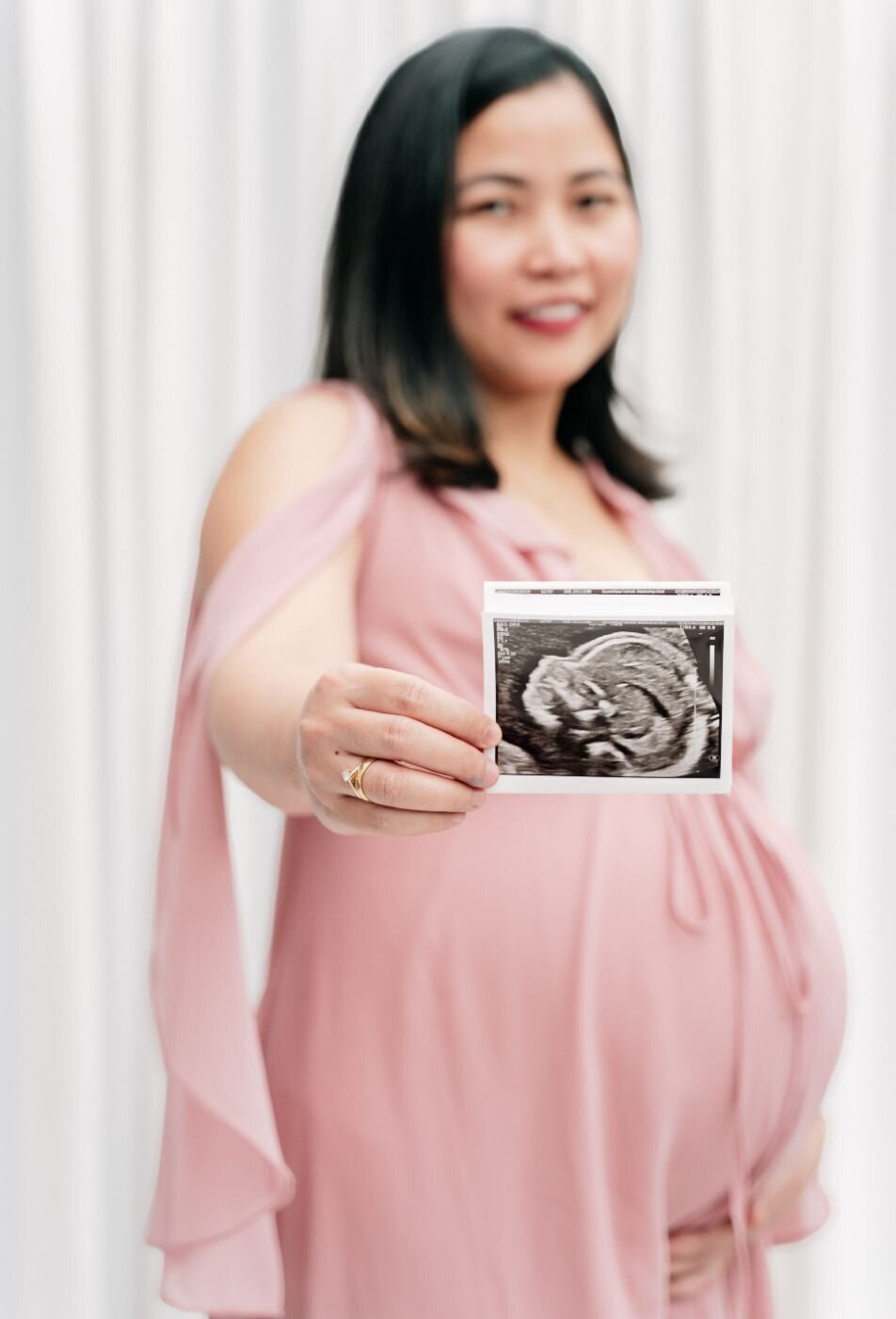 A close-up, color photo of an ultrasound picture being held by two parents. The ultrasound shows a clear profile view of the baby in the womb. The parents' hands, with a wedding ring visible on the woman's finger, are gently holding the image. The background is a blurred, warm-toned environment.