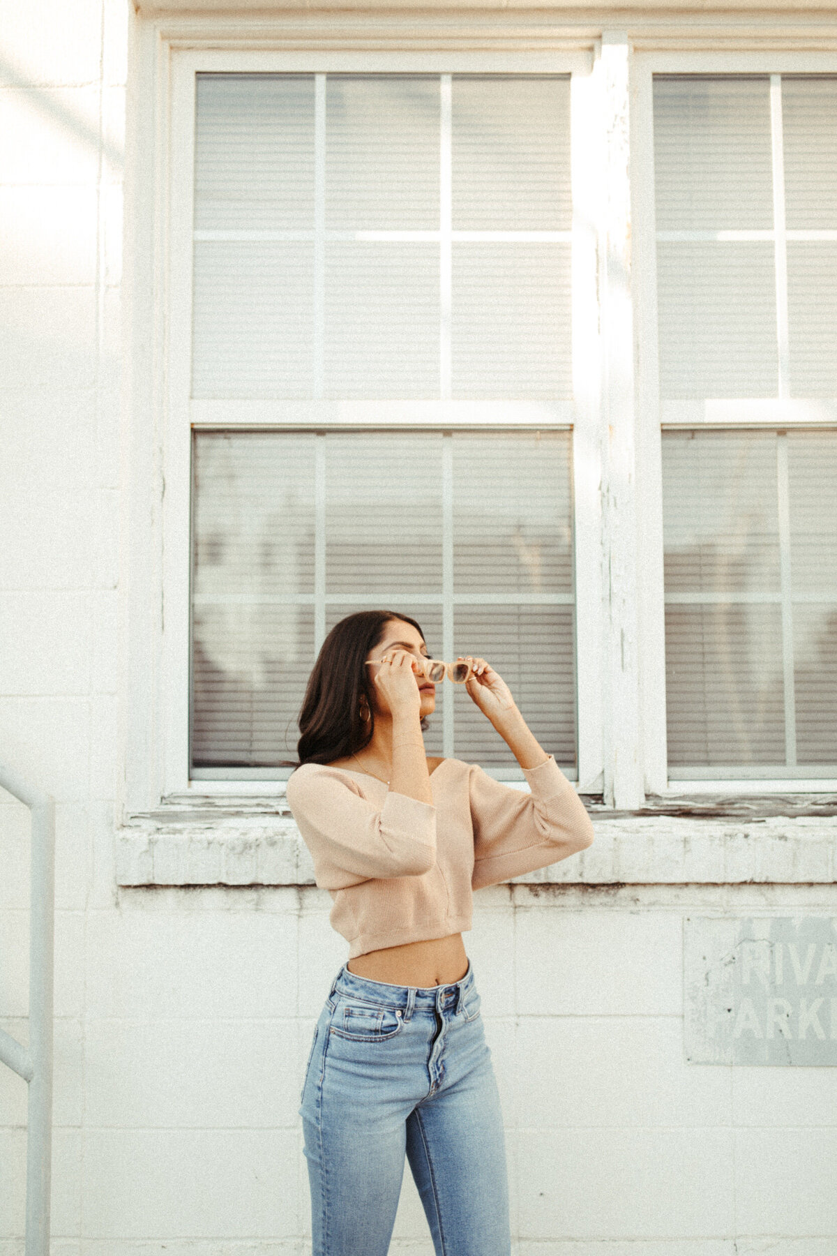 Senior girl in crop top putting on vintage sunglasses in front of window on a building downtown