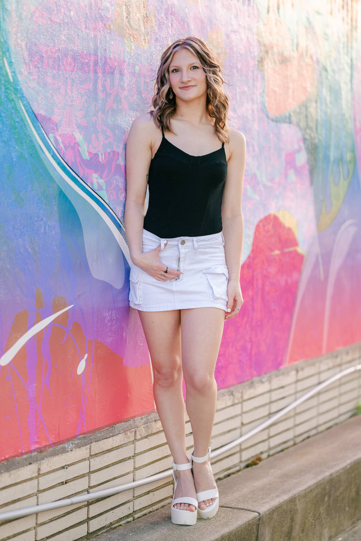 A high school senior, wearing a black top and white skirt and heels leans against a colorful mural. She is popping one leg and has her hand hooked through her belt loops. She is at her senior session with Chesapeake senior photographer Justine Renee Photography.