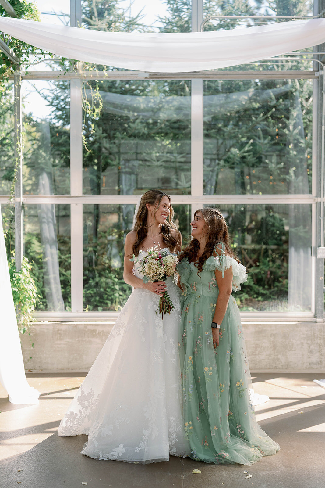 Bride and her mom smiling together inside the greenhouse at The Ivy House during family wedding portraits.