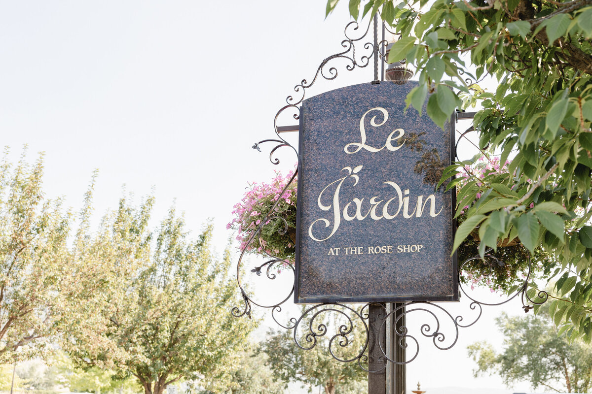 An elegant wrought iron sign reading "Le Jardin at The Rose Shop" is adorned with hanging pink flowers, set against a bright blue sky and lush green trees.