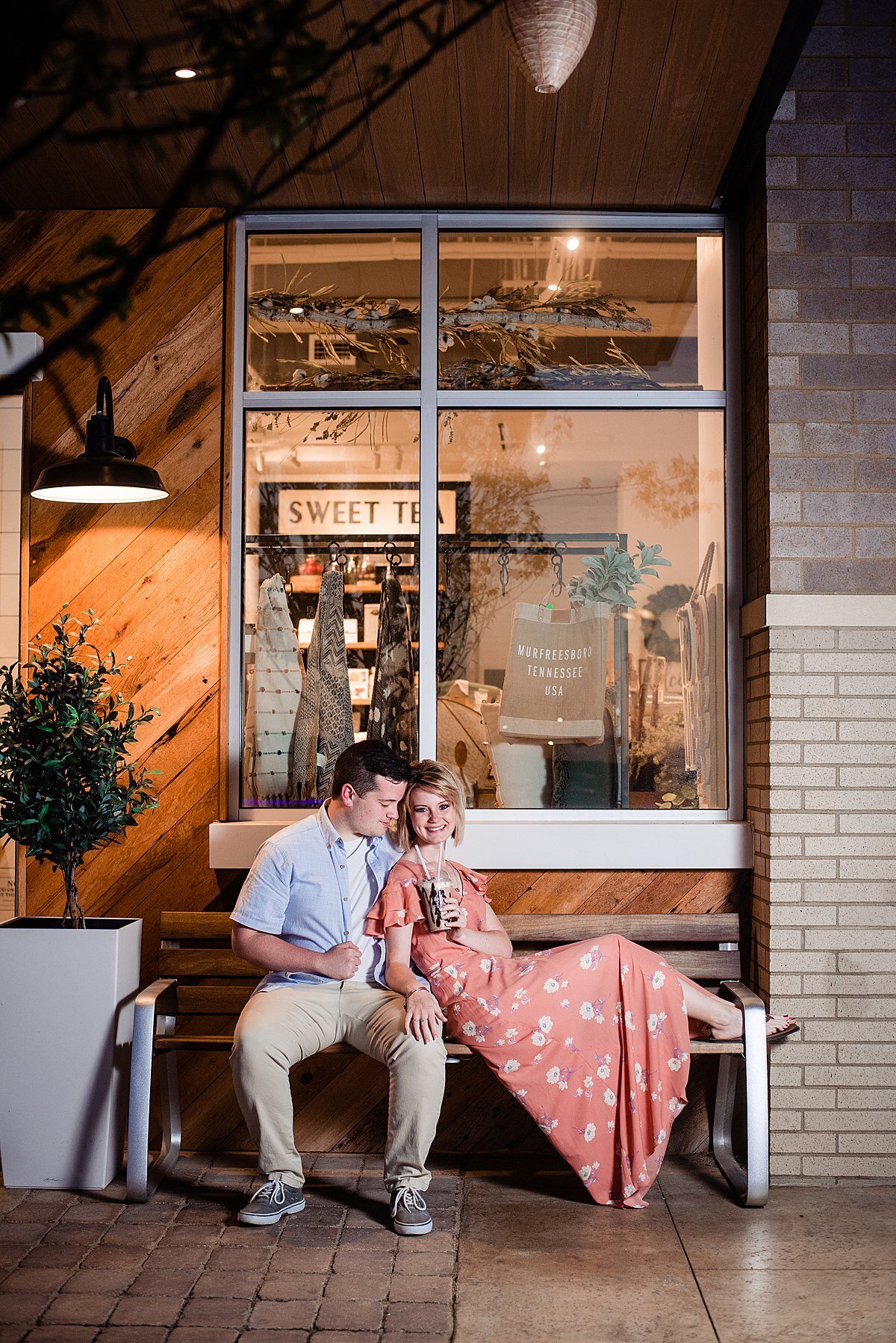 The bride and groom sit on a metal park bench in front of a large glass store window with a sweet tea sign. The  groom sits on the bench with his feet on the ground looking over at the bride. He is wearing khaki pants, a white t shirt and a blue button down shirt. The bride sits sideways with her back against the groom's chest and her feet propped up and she sips on a soda from a glass with a straw. She smiles at the camera. The bride is wearing a peach dress with a white floral pattern,