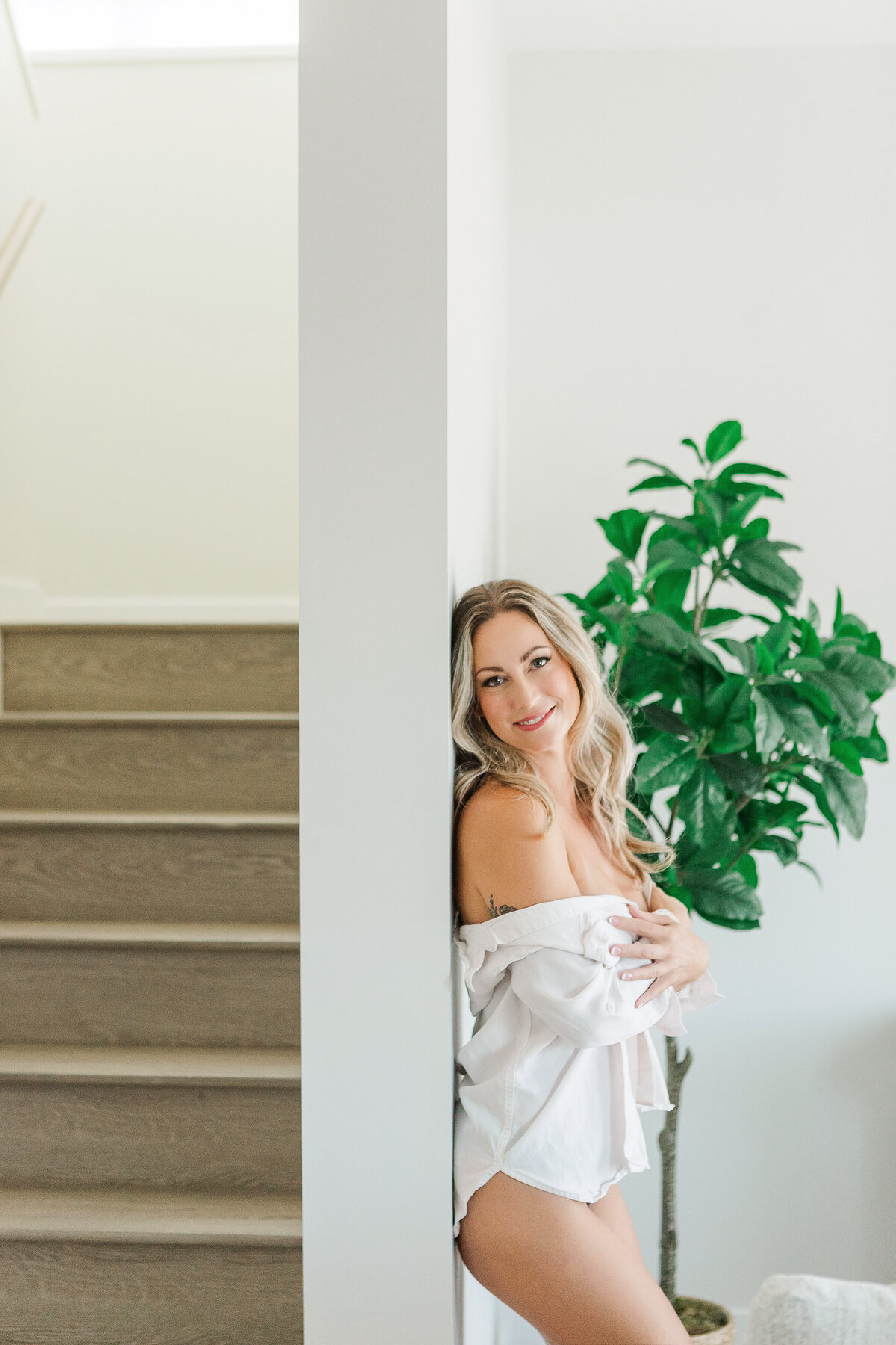 A woman leaning against the wall near a staircase, smiling warmly while wearing an oversized shirt in a bright, airy space.