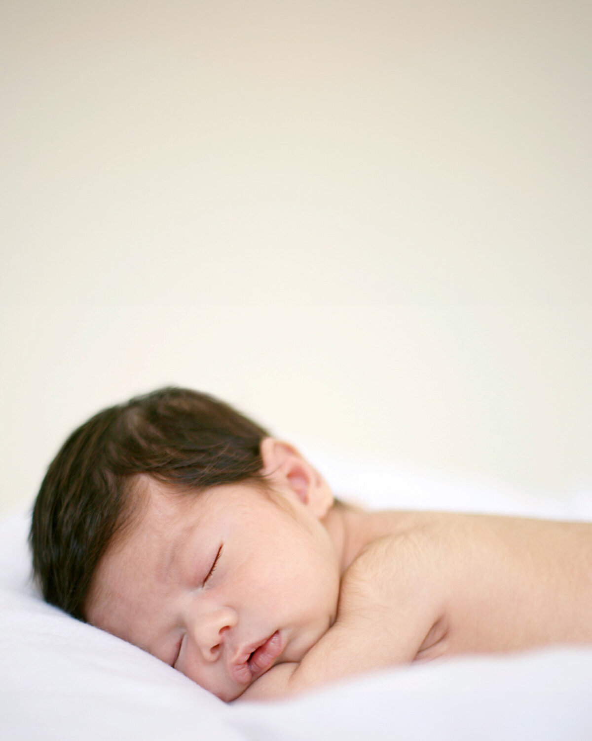 Newborn baby with full head of hair sleeping on stomach with hands tucked under chin on white bed, natural portrait by Menlo Park newborn photographer.