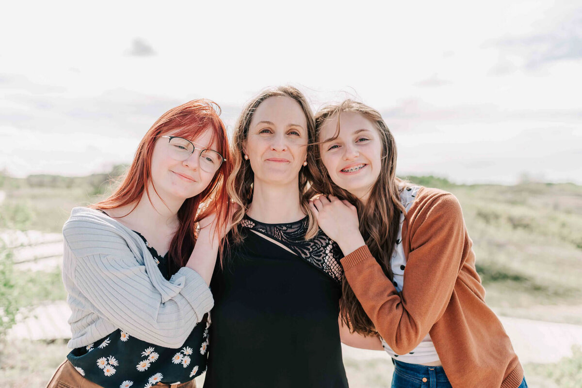 2 teenage daughters stand close to their mom and lean on her shoulders. 