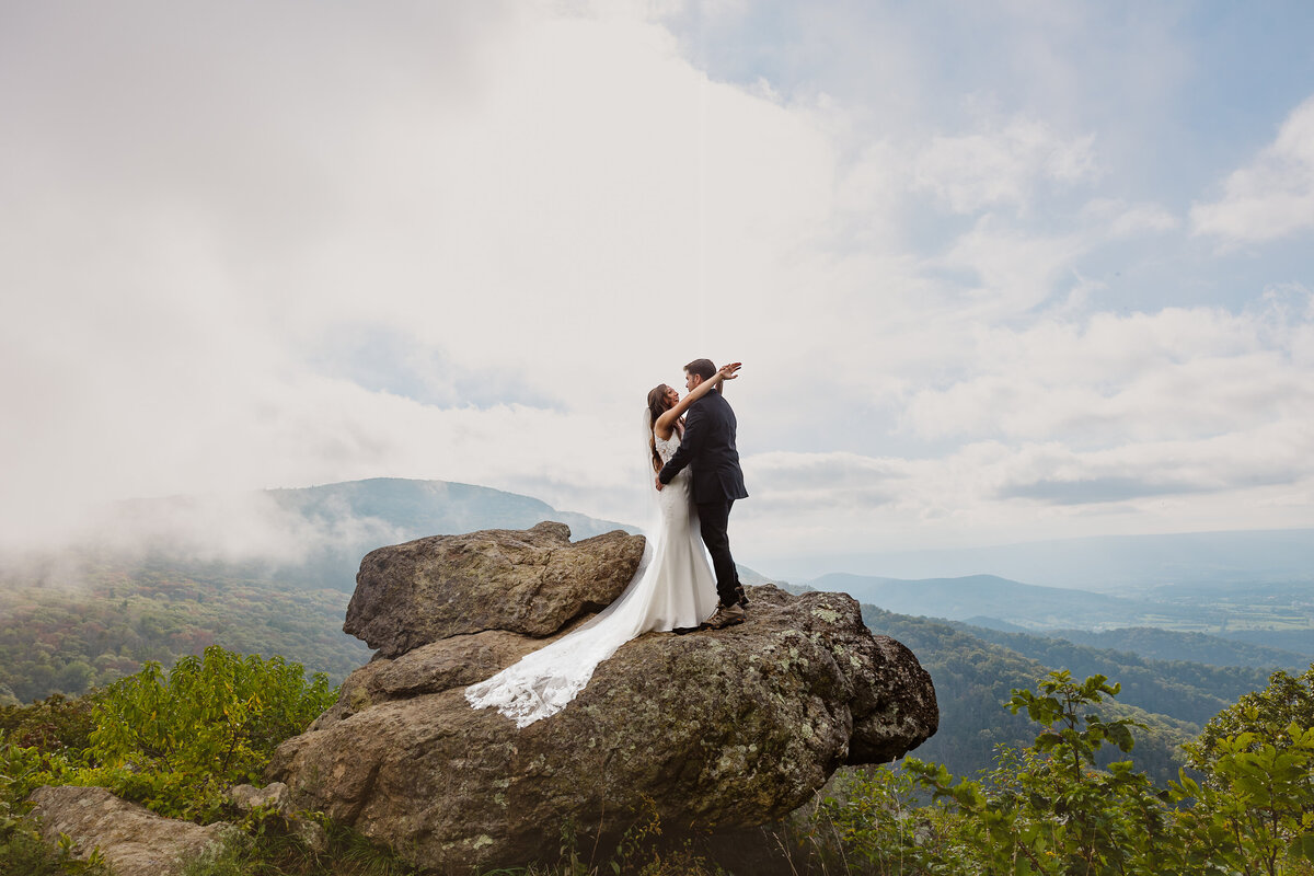 A couple stands on a rock at an overlook at Shenandoh National Park. The bride is facing the groom, and has her arms wrapped around the back of his neck.