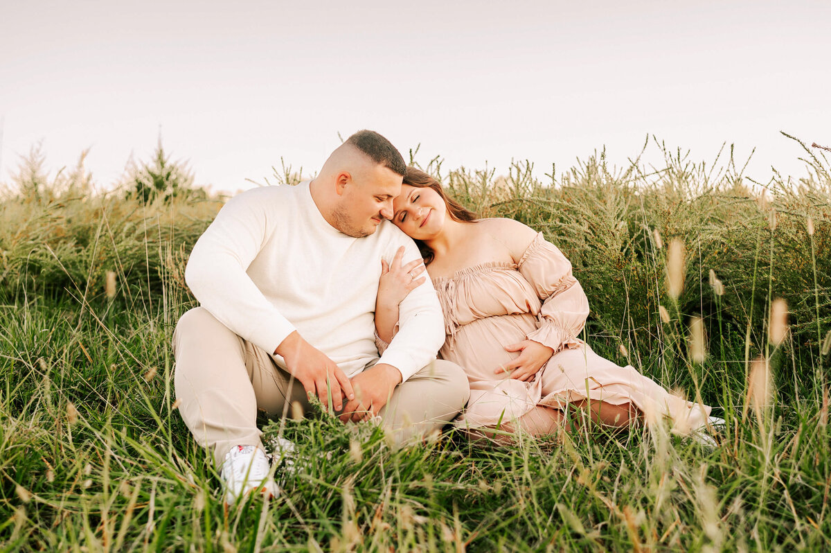 pregnant mom leaning on husbands shoulder sitting in grass at sunset during Joplin MO maternity photography session