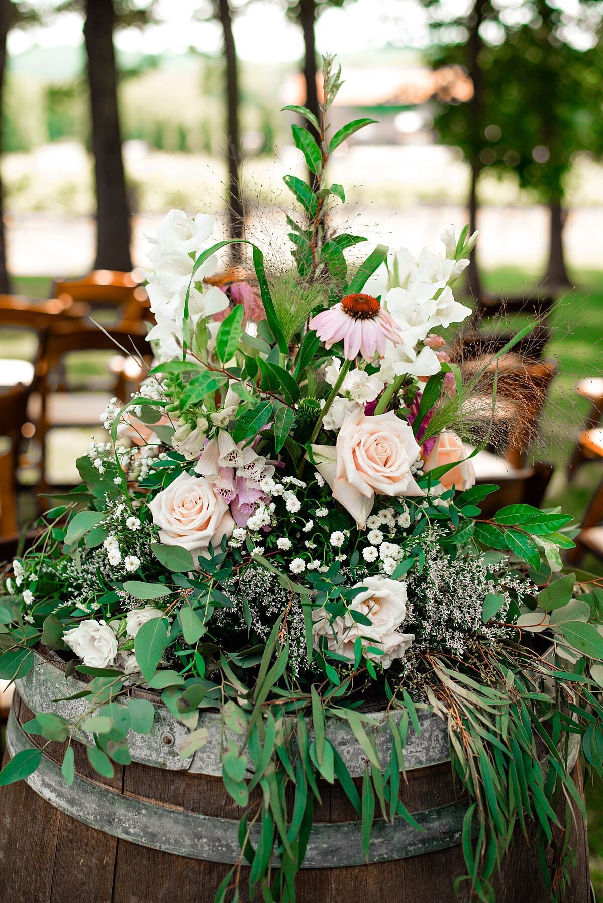 Sitting on top of a wine barrel a large cascading floral arrangement of assorted greenery with pink roses, pink hyacinth, pink carnations, pink cosmos and baby's breath at Arrington Vineyards garden party
