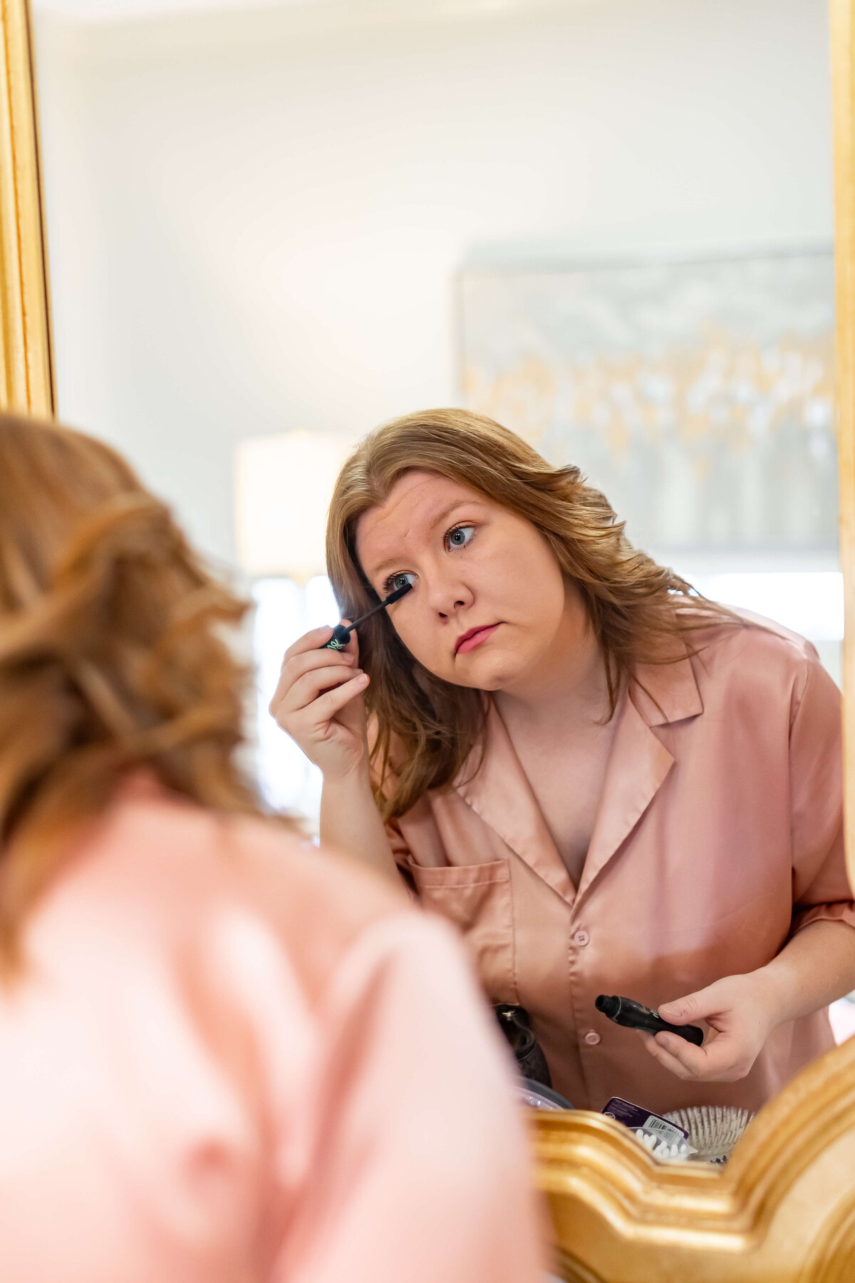 A woman in a pink satin pajama top applies mascara while looking into a mirror. Her hair is styled in loose waves, and she appears focused on her reflection.