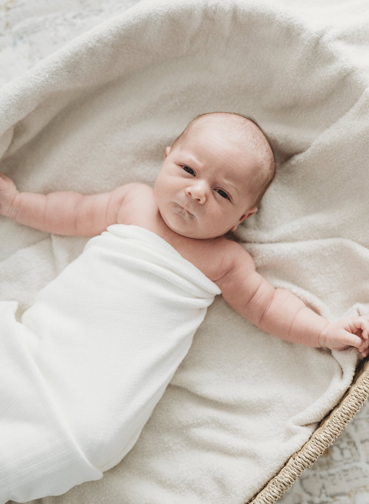 Newborn baby wrapped in a white swaddle, lying in a basket with a soft smile during a Kansas City newborn session.