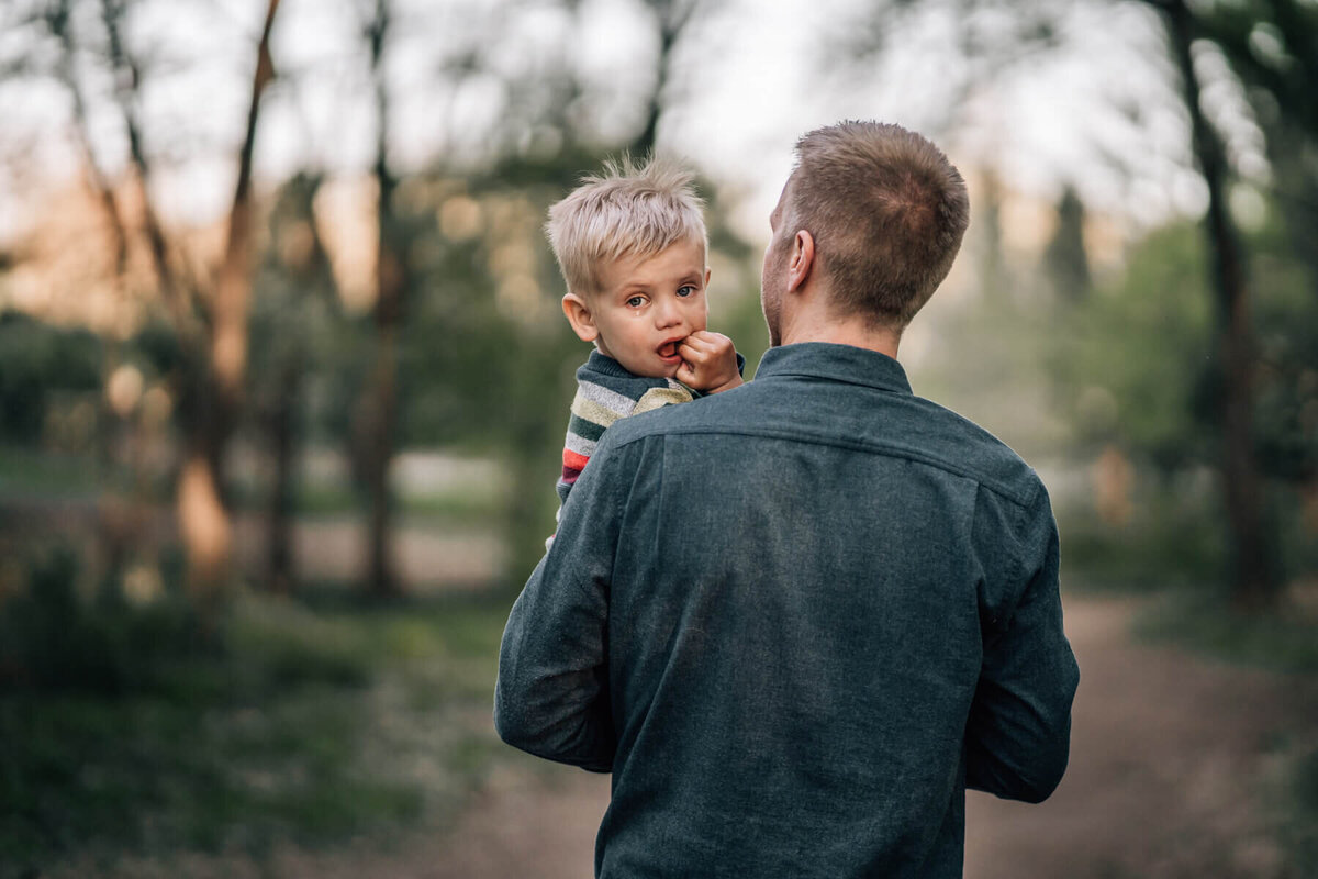 A large tear rests on a little boy's cheek as he looks back over his father's shoulder.