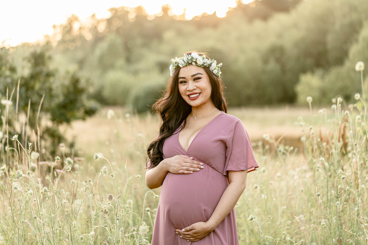 brown-skinned pregnant woman in mauve dress standing in tall grasses wearing a flower crown at maternity photography session.