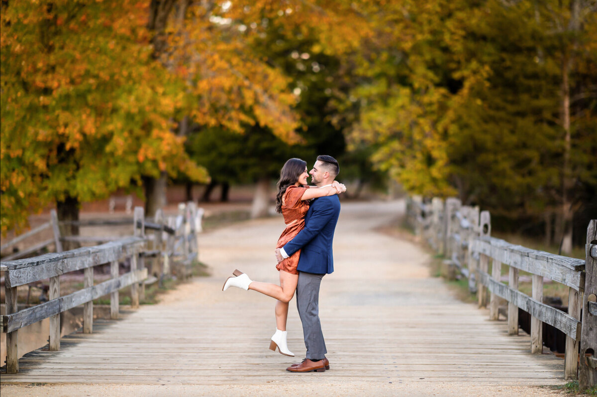Woman in a orange dress and man and a blue jacket, posing on a bridge, and that Stowe Village in the fall for their engagement session