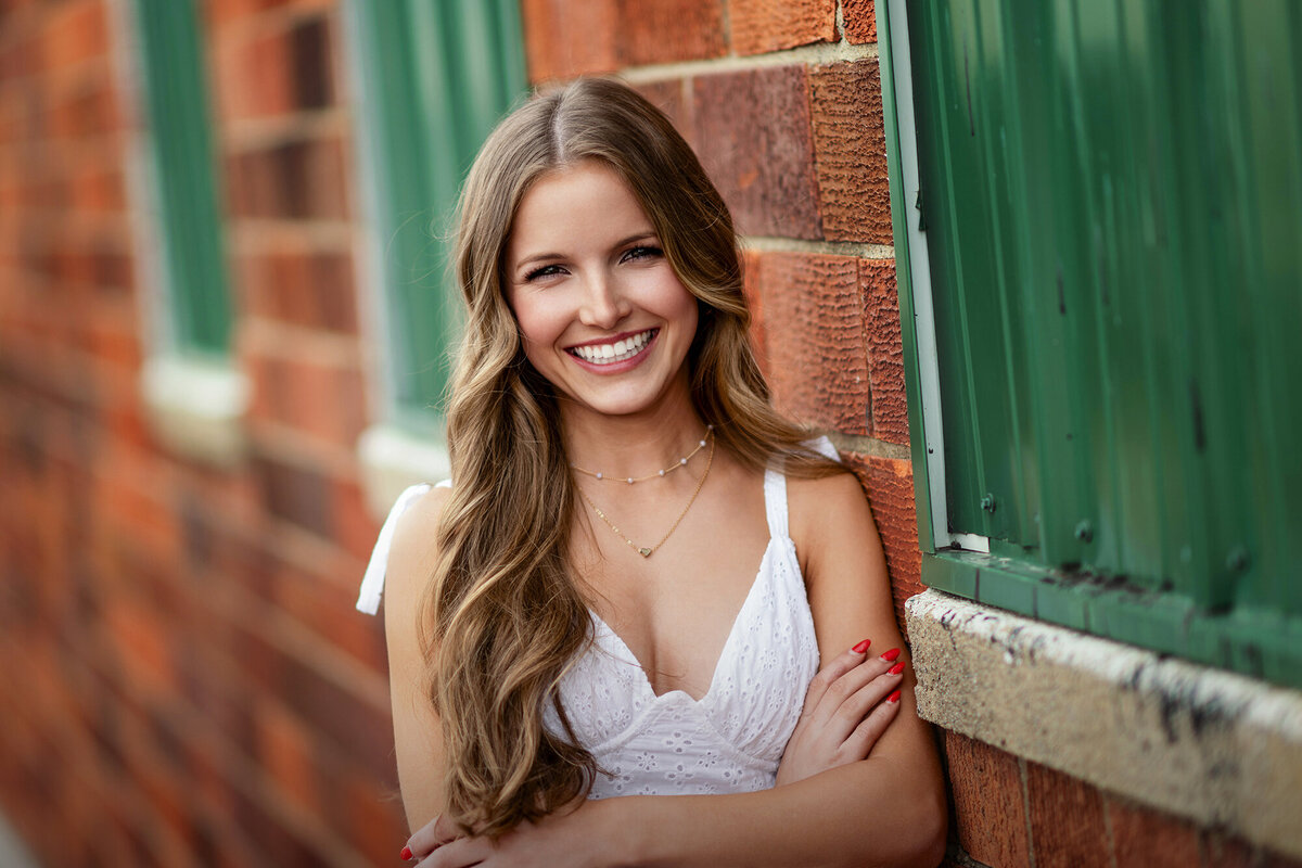 A high school senior in a white dress leans on a brick wall while smiling