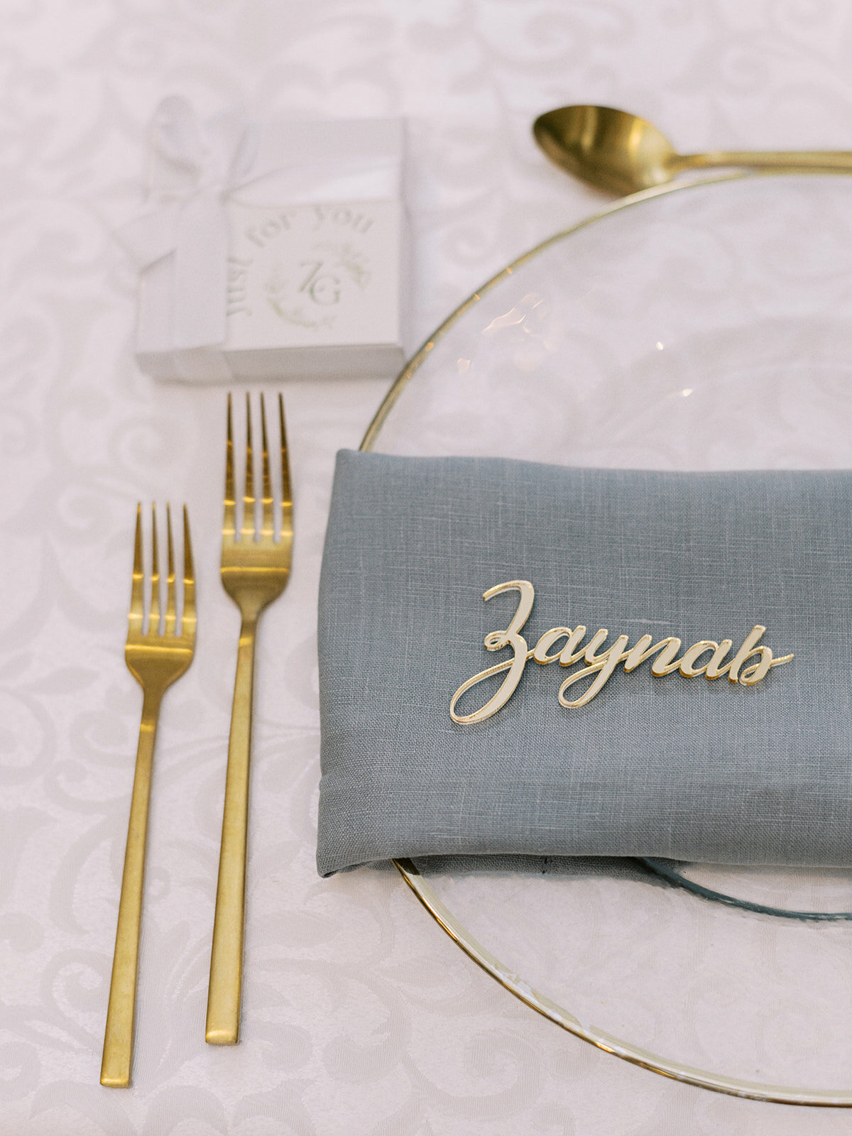 A classic Calgary wedding place setting featuring a blue napkin with the name "Zaynab" in gold lettering, two gold forks, a small gift box, and a clear plate on a white tablecloth. Perfect for an elegant Wedding at Fairmont Palliser Calgary.