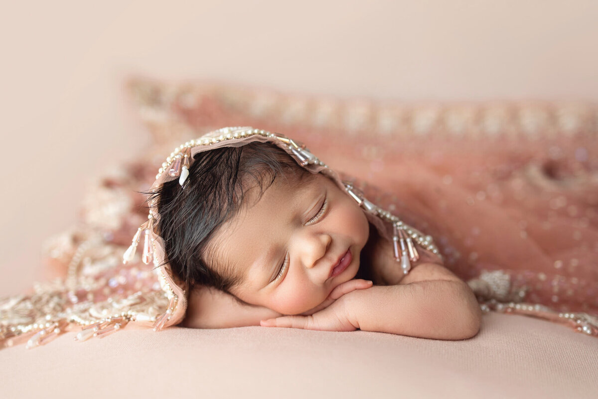 Newborn baby girl with dark hair, wrapped in a delicate peach lace fabric, sleeping peacefully on a matching peach blanket.