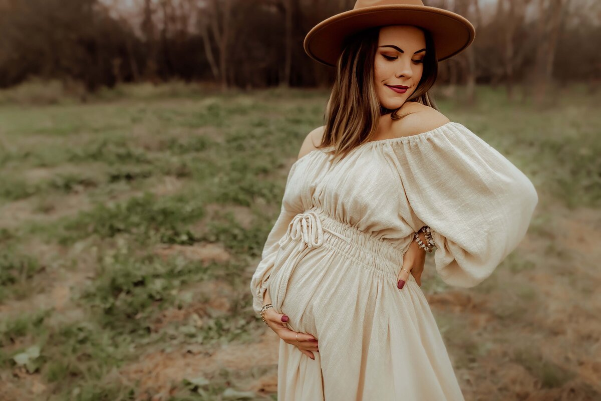A person in a beige off-shoulder dress and brown hat stands outdoors, holding their pregnant belly with one hand and resting the other hand on their hip. Trees are visible in the background.