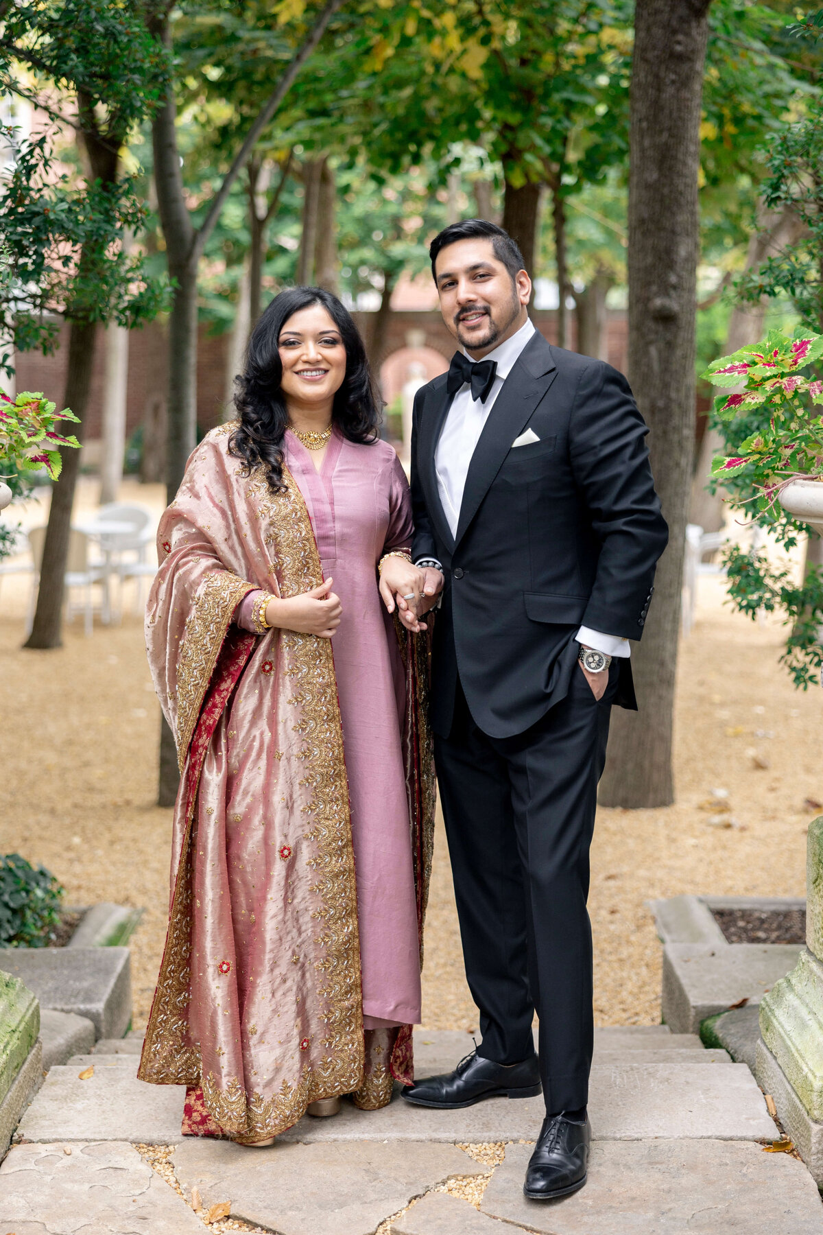 A smiling couple stands arm in arm outdoors. The woman wears a traditional pink and gold embroidered gown, while the man wears a black tuxedo. They are surrounded by greenery and stone steps.