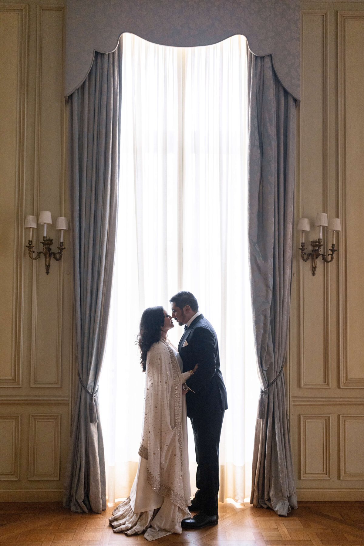 A couple stands facing each other in an elegant room, silhouetted against large, softly lit windows with long curtains. The woman wears a flowing white dress, while the man is in a dark suit. The scene is serene and intimate.