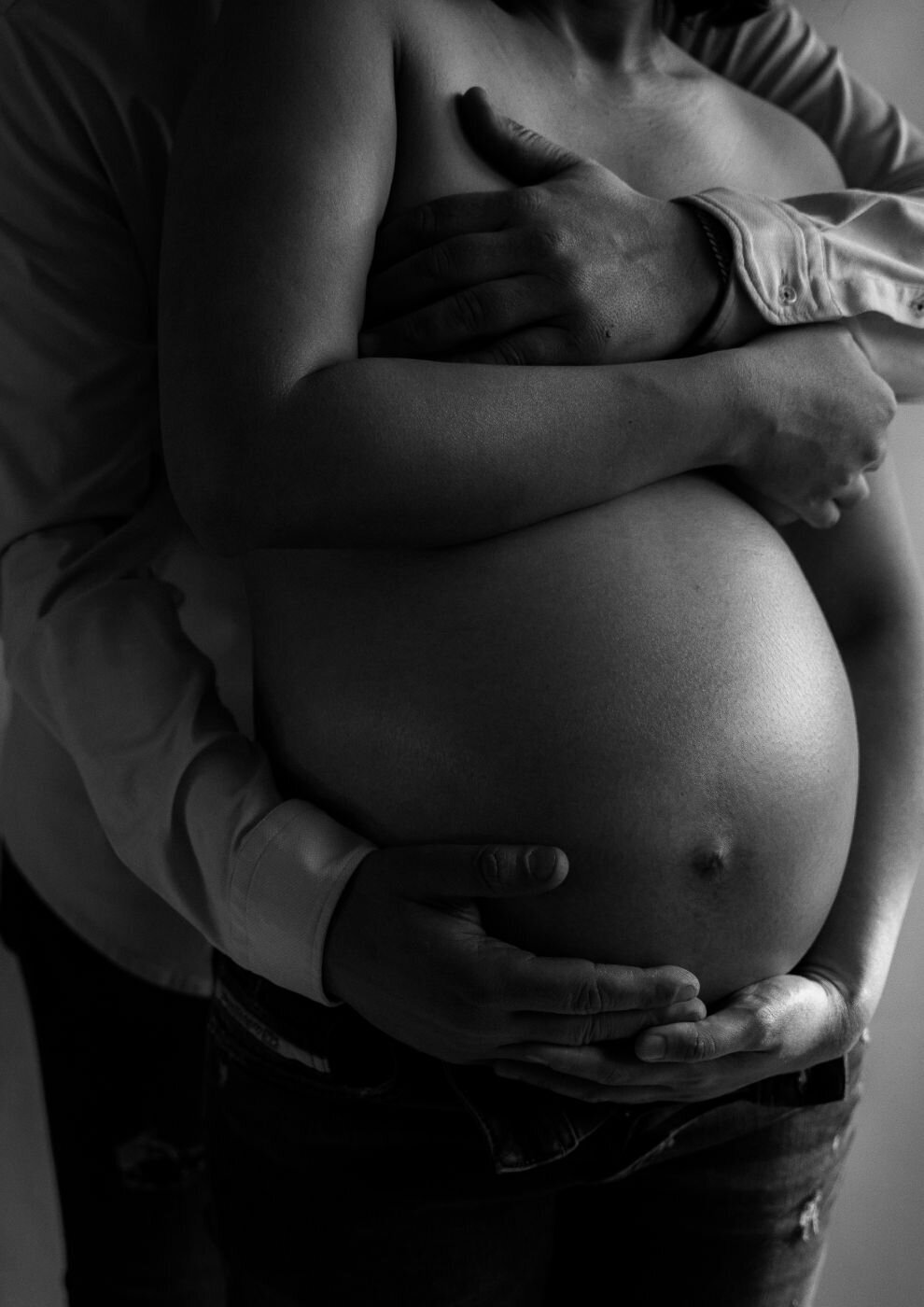 A black and white portrait of an expecting couple. The man stands behind the pregnant woman, gently holding her baby bump with his hands. The woman's head is tilted back to rest against his shoulder, and she is holding her hands on her stomach as well. They are both looking down at her belly with loving expressions.