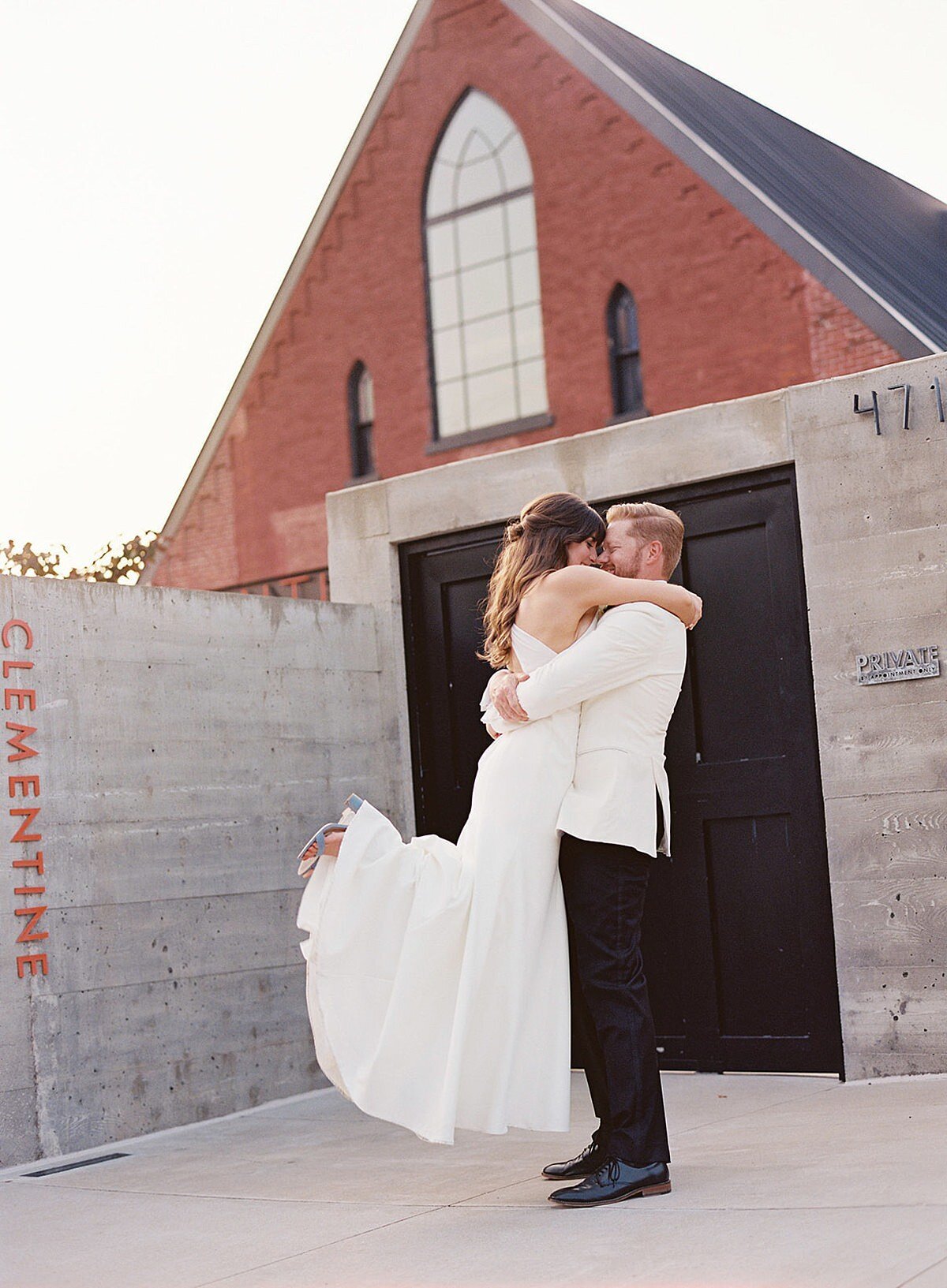 The bride, a brunette, wearing a silk halter top sheath dress embraces the groom who is wearing a tuxedo with a white jacket as he lifts her up off the ground and twirls her around. The gray walls of the entry of Clementine Hall are accented with the word Clementine written in orange. The exposed red brick pediment of the building rises above them.