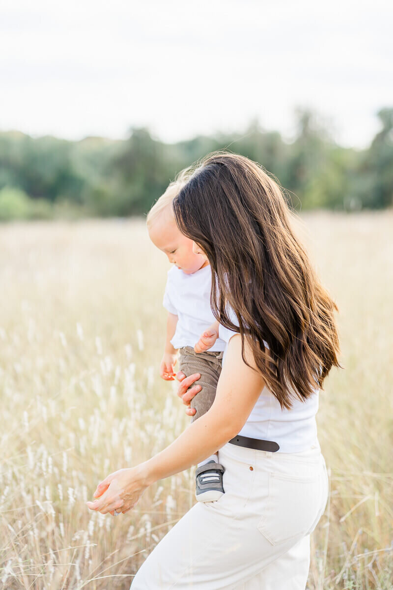 a mother holding her toddler son walks in a field of tall grass for their family photos.