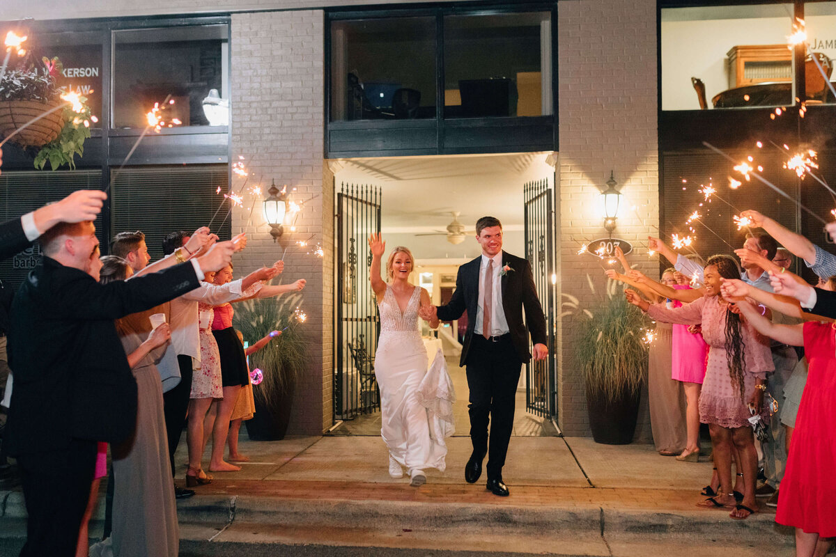 A bride and groom smile and walk hand in hand outside a building at night, surrounded by guests holding lit sparklers and celebrating their wedding.