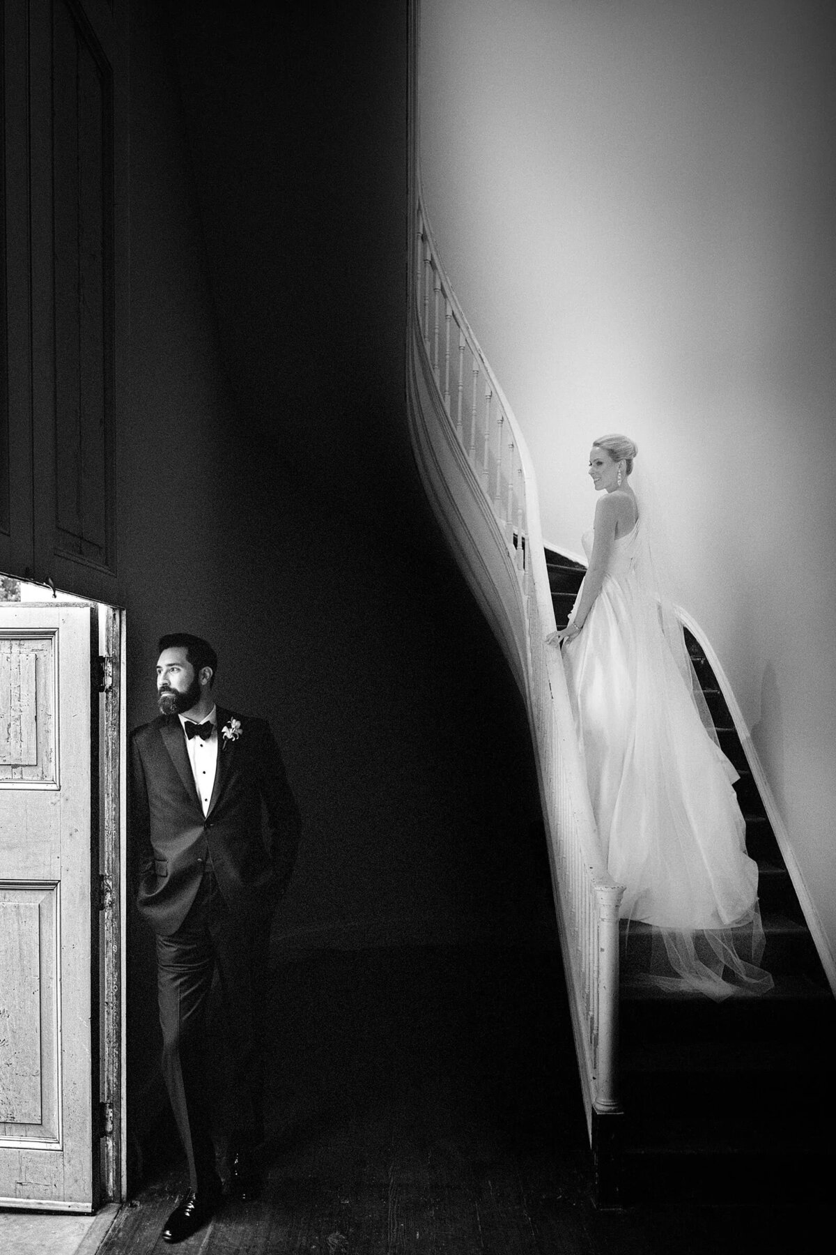 A dramatic black and white photo of a bride ascending a staircase while the groom waits below