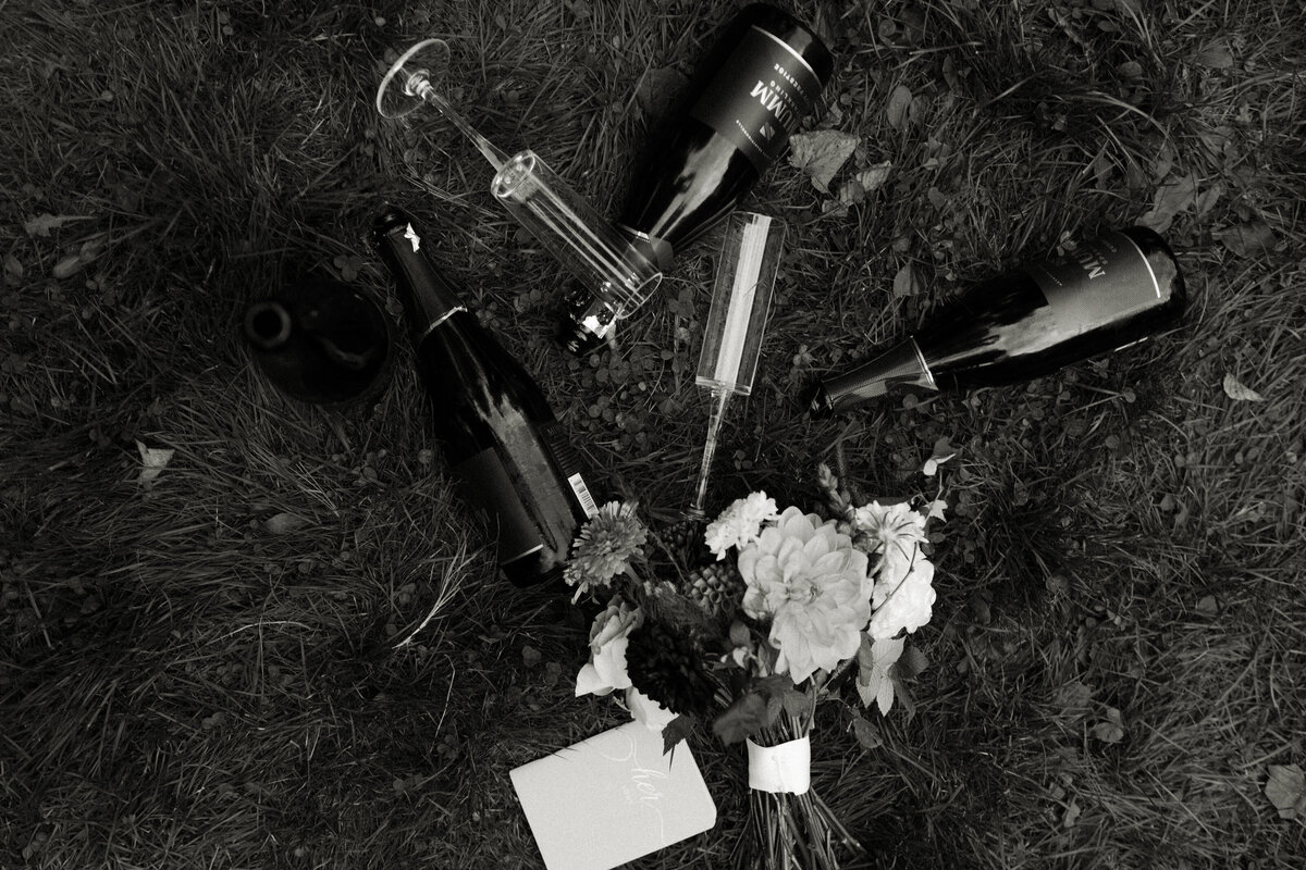 Champagne, champagne flutes, and a bridal bouquet rest on the ground among fall foliage.