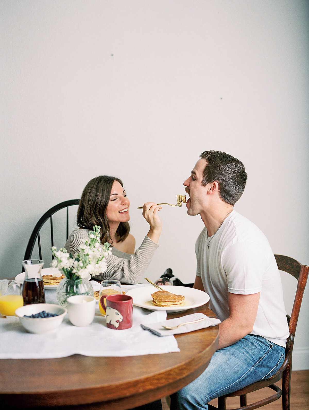 Woman feeding man a bite of food at a table with breakfast on it