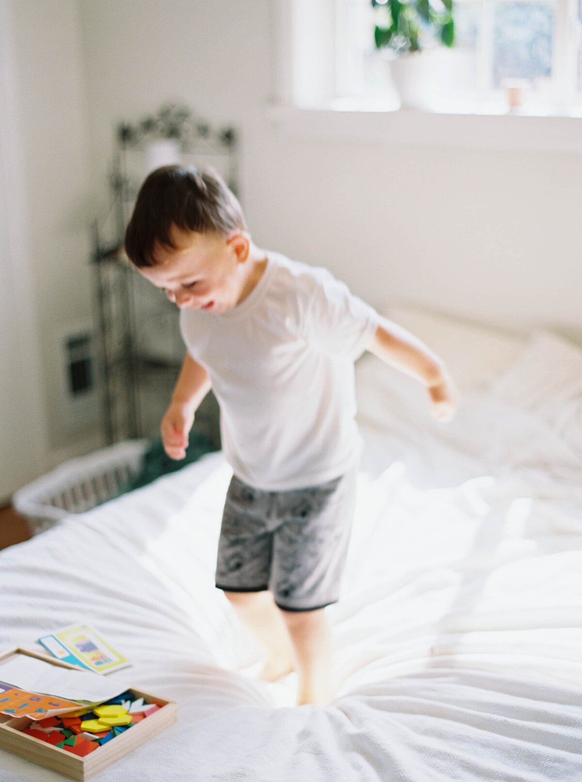 Photo of a young boy jumping on a bed inside a brightly lit room with big windows and white sheets.