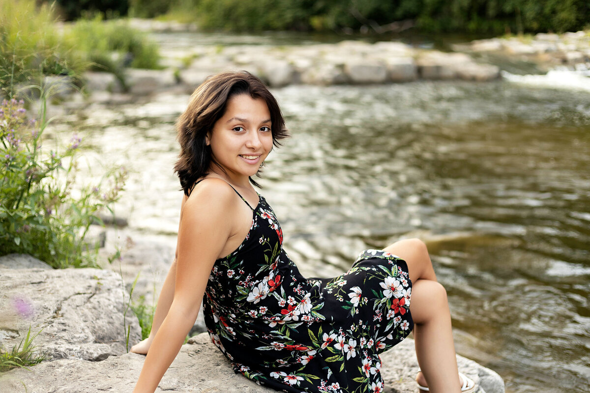 Ann Arbor Argo Park Canoe & Kayak Senior Photoshoot | Ann Arbor Michigan Senior Photographer, Senior girl with black hair in black floral dress sitting on rocks by the Huron River at sunset