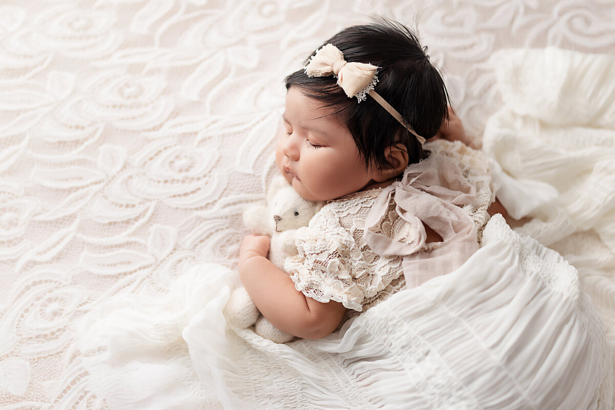 Sleeping newborn baby girl with dark hair lying on a lace blanket, wearing a vintage cream outfit and bow headband, cuddling a small teddy bear.