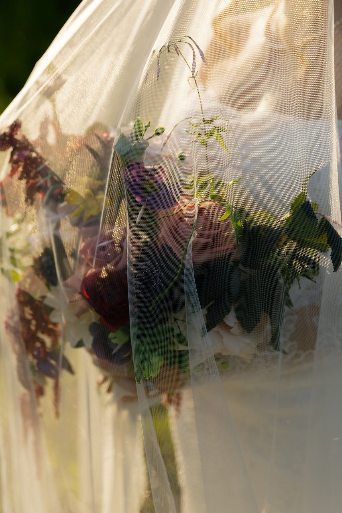 Soft and romantic bridal bouquet photographed through a delicate veil at sunset, highlighting mauve roses, burgundy blooms, purple floral accents, and natural greenery, arranged in a textured, organic style by a wedding florist.