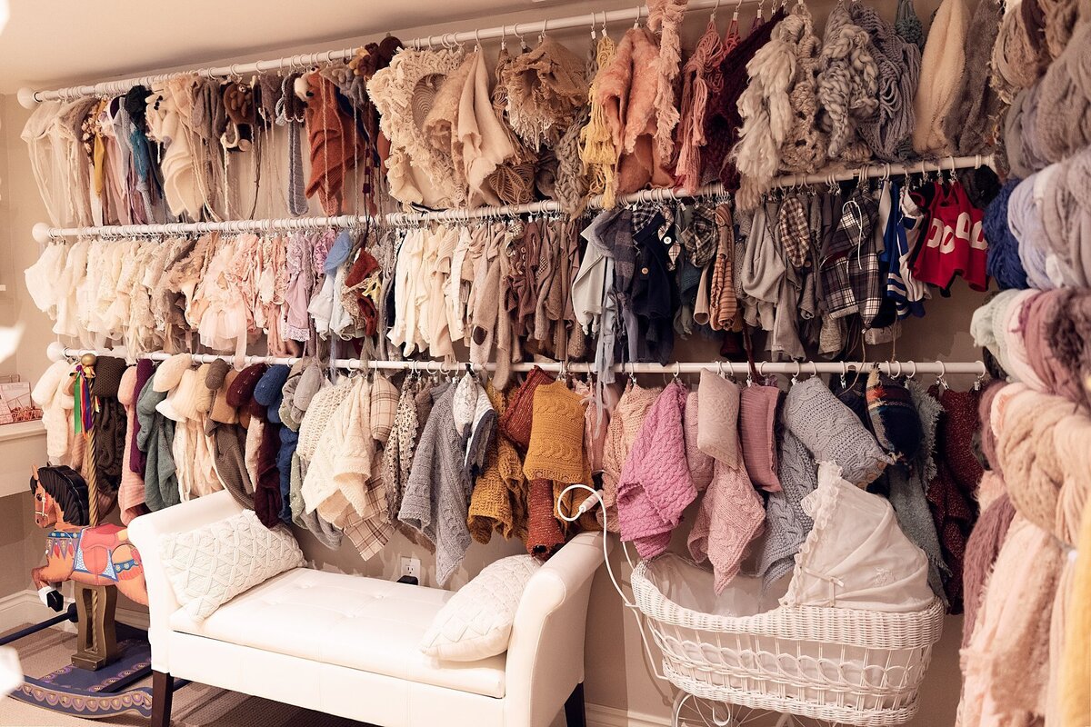 Large wall of hanging newborn photography outfits and wraps arranged by colour inside an Oakville studio wardrobe room.
