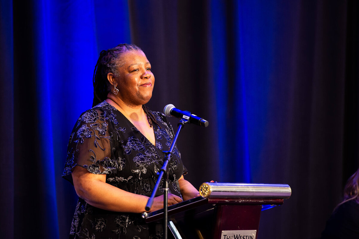A black woman in a blue dress speaking into a microphone addressing attendees during a 2-day corporate conference at the Westin Hotel.  Captured by Ottawa Event Photographer JEMMAN Photography COMMERCIAL