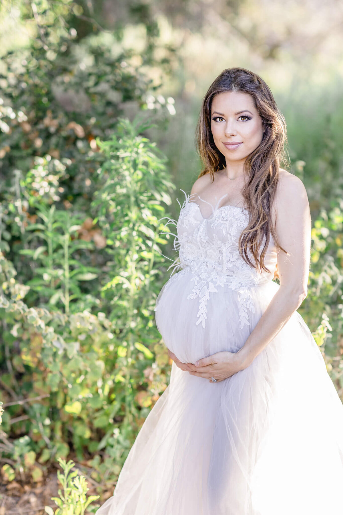 A pregnant woman in a white lace and tulle dress stands outdoors, surrounded by greenery. She cradles her belly with both hands and looks at the camera, with sunlight filtering through the leaves.
